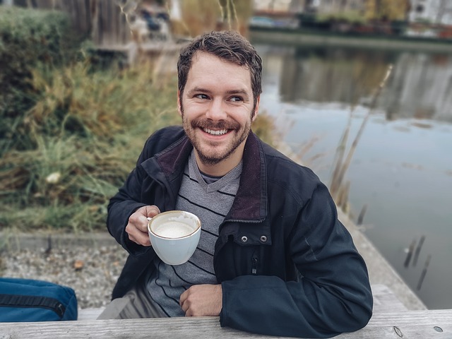 Usmiaty muž v čiernej bunde sedí pri stole vedľa jazera a pije kávu
